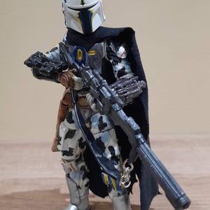 May include: A detailed action figure of a character in a white and blue helmet, black cape, and patterned armor. The figure is holding a large black rifle. The figure has silver boots and a brown holster. The overall theme is science fiction.