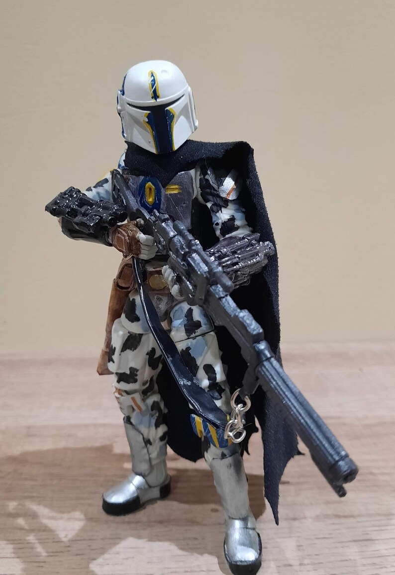 May include: A detailed action figure of a character in a white and blue helmet, black cape, and patterned armor. The figure is holding a large black rifle. The figure has silver boots and a brown holster. The overall theme is science fiction.