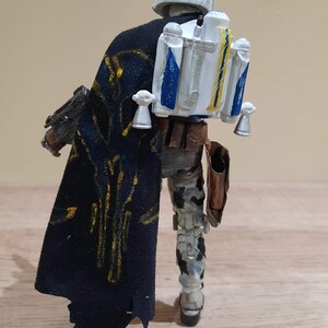 May include: A detailed action figure of a character from a science fiction franchise, viewed from the back. The figure wears a white helmet with blue accents, a black cape with gold detailing, and a jetpack with blue and yellow stripes. The figure is on a wooden surface.