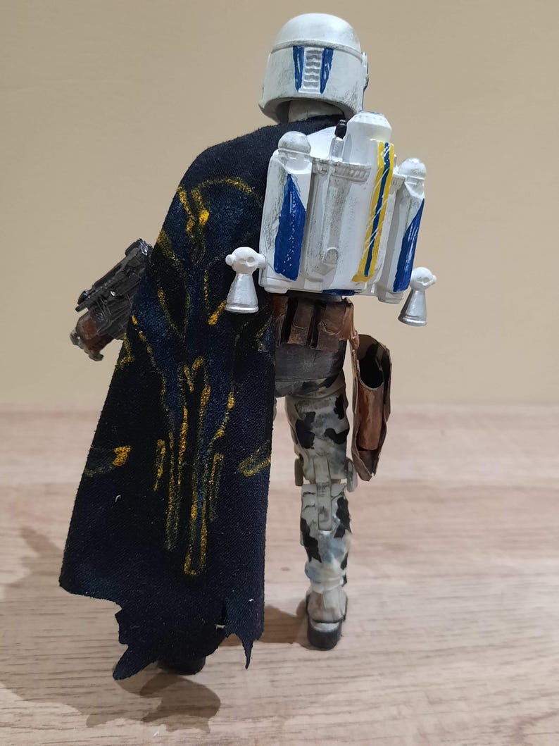 May include: A detailed action figure of a character from a science fiction franchise, viewed from the back. The figure wears a white helmet with blue accents, a black cape with gold detailing, and a jetpack with blue and yellow stripes. The figure is on a wooden surface.