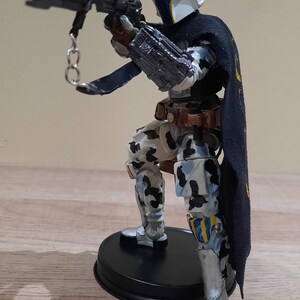 May include: A detailed action figure of a character in a white helmet with blue and yellow accents, holding a black weapon. The figure wears a black cape with gold accents and camouflage-patterned armor. It stands on a black base.