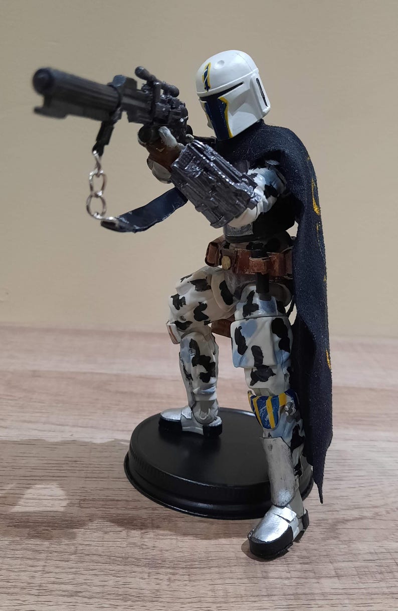 May include: A detailed action figure of a character in a white helmet with blue and yellow accents, holding a black weapon. The figure wears a black cape with gold accents and camouflage-patterned armor. It stands on a black base.