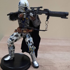 May include: A detailed action figure of a character in a white and blue helmet and armor, holding a large black rifle. The figure has a black cape and is posed on a black circular base. The figure's armor has a camouflage pattern.