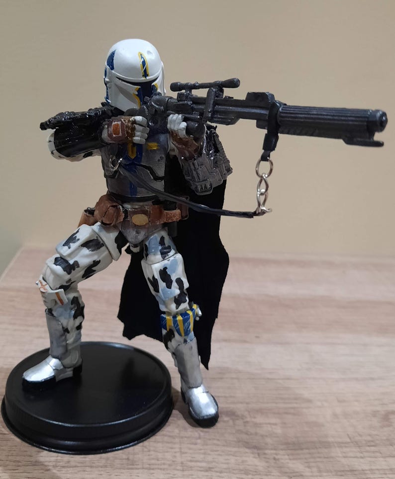May include: A detailed action figure of a character in a white and blue helmet and armor, holding a large black rifle. The figure has a black cape and is posed on a black circular base. The figure's armor has a camouflage pattern.