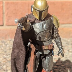 K&ouml;nnte beinhalten: Detaillierte Actionfigur eines Charakters mit goldenem Helm und dunkler R&uuml;stung, der eine Waffe h&auml;lt. Die Figur hat einen braunen Umhang und posiert vor einer Backsteinmauer. Die Figur ist etwa 15 cm gro&szlig;.