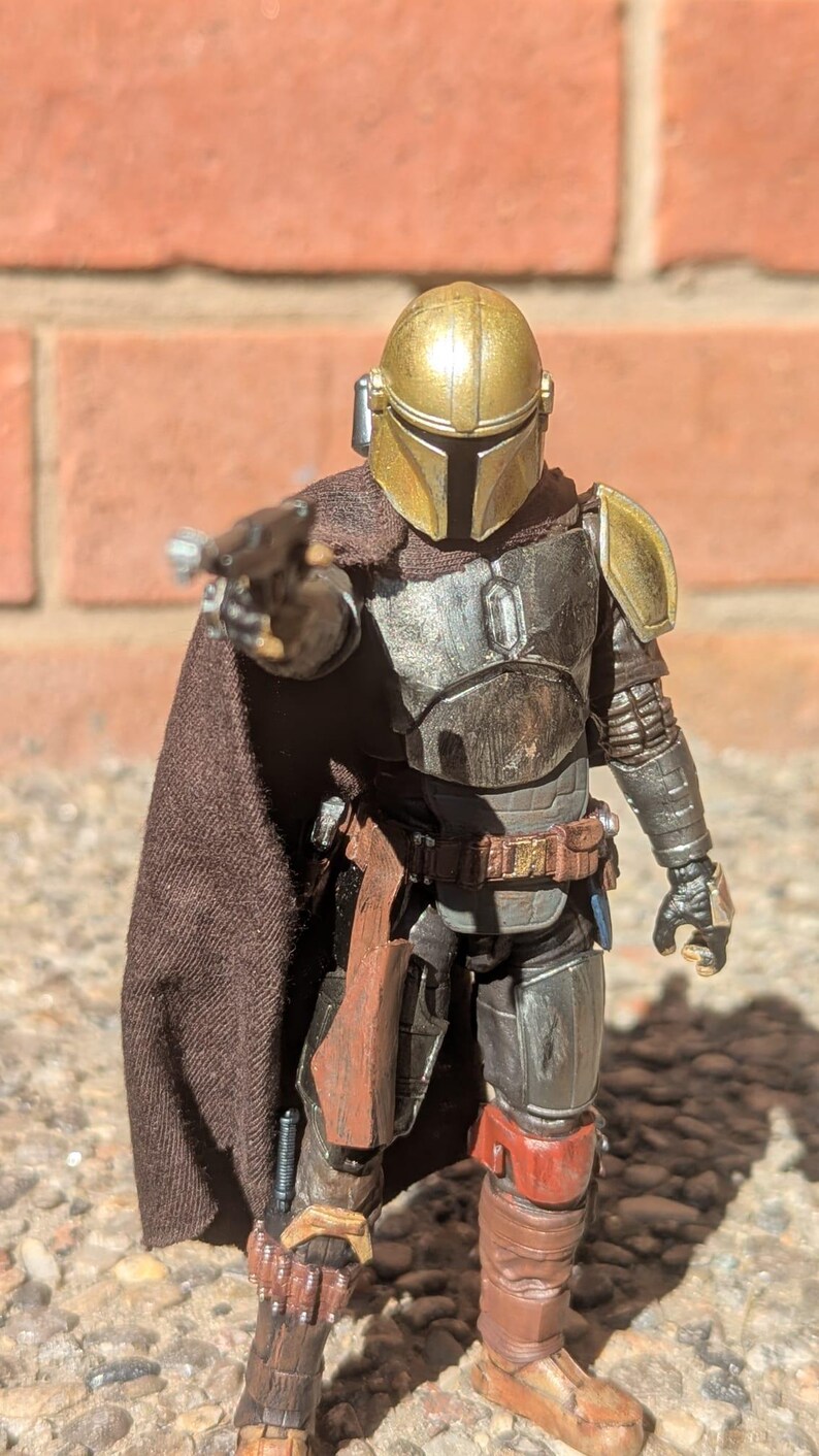 K&ouml;nnte beinhalten: Detaillierte Actionfigur eines Charakters mit goldenem Helm und dunkler R&uuml;stung, der eine Waffe h&auml;lt. Die Figur hat einen braunen Umhang und posiert vor einer Backsteinmauer. Die Figur ist etwa 15 cm gro&szlig;.
