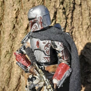K&ouml;nnte beinhalten: Eine silberne und rote Actionfigur eines Mandalorianer-Kriegers mit einem schwarzen Umhang und einem Blastergewehr. Die Figur ist detailliert und hat einen verwitterten Look.