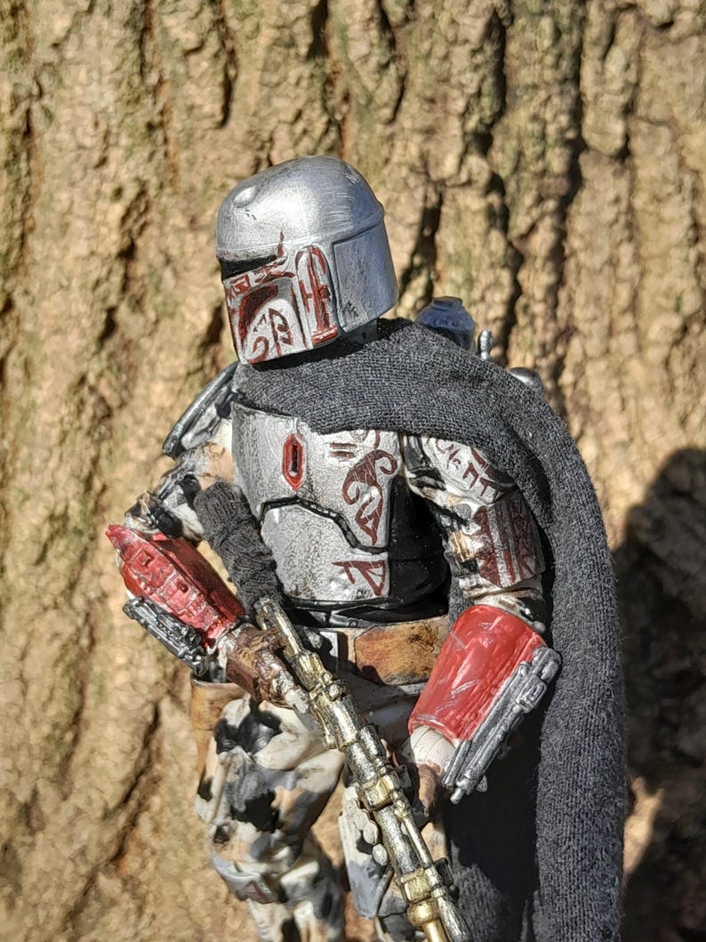 K&ouml;nnte beinhalten: Eine silberne und rote Actionfigur eines Mandalorianer-Kriegers mit einem schwarzen Umhang und einem Blastergewehr. Die Figur ist detailliert und hat einen verwitterten Look.