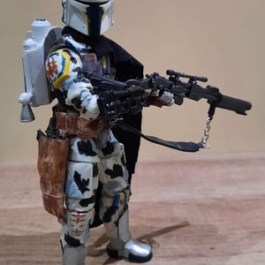 May include: A detailed action figure of a character in a white, blue, and yellow helmet and armor, holding a black rifle. The figure has a jetpack and a brown holster. The figure is standing on a wooden surface.