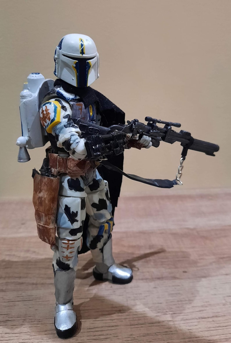 May include: A detailed action figure of a character in a white, blue, and yellow helmet and armor, holding a black rifle. The figure has a jetpack and a brown holster. The figure is standing on a wooden surface.