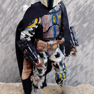 May include: A detailed action figure of a Mandalorian warrior. The figure is clad in weathered armor with a black cape. The helmet is white with blue and yellow accents. The figure is posed on a textured surface.