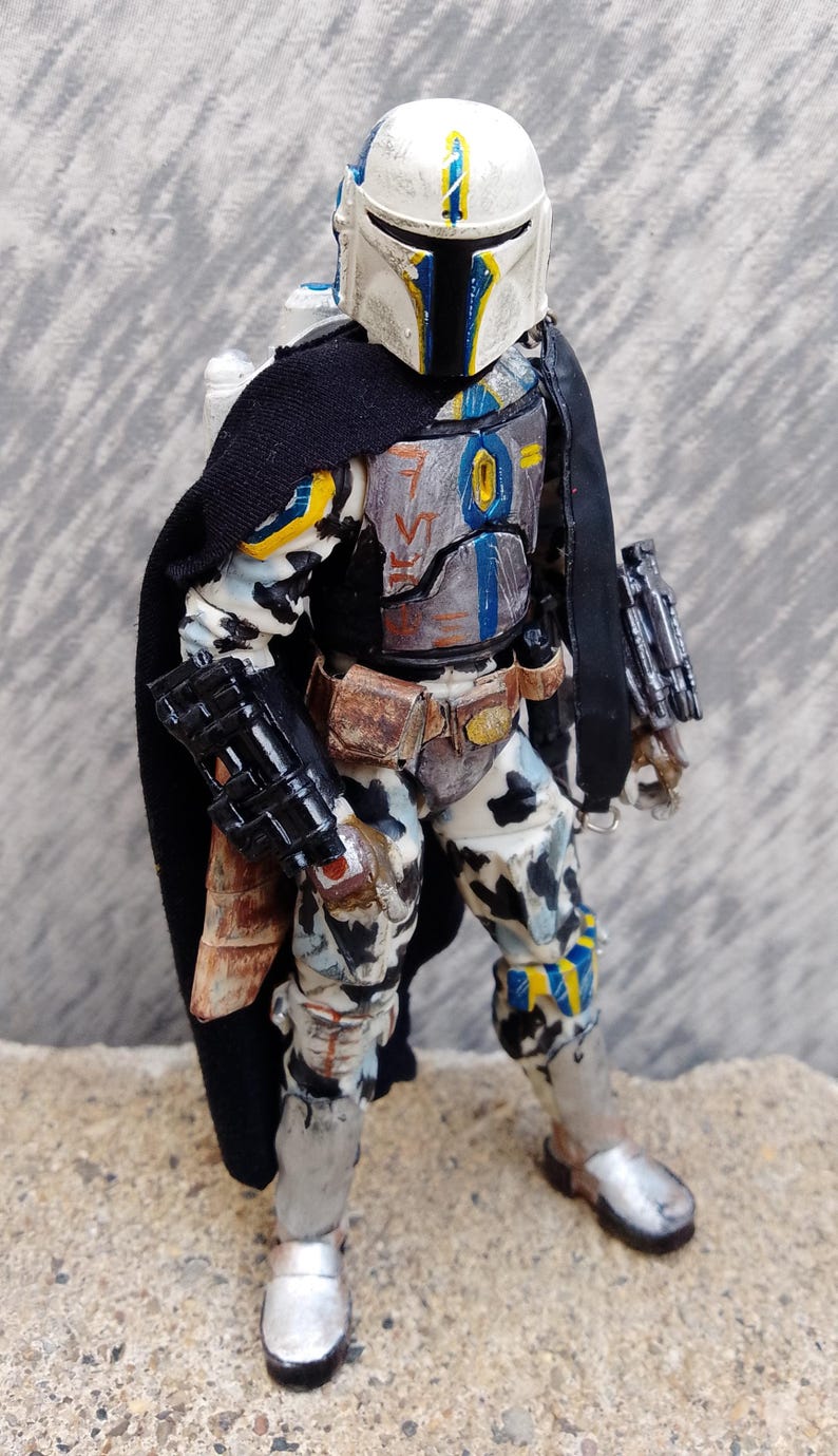 May include: A detailed action figure of a Mandalorian warrior. The figure is clad in weathered armor with a black cape. The helmet is white with blue and yellow accents. The figure is posed on a textured surface.
