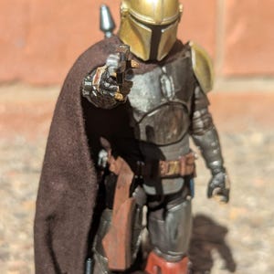 K&ouml;nnte beinhalten: Detaillierte Actionfigur eines Charakters in metallischer R&uuml;stung, mit goldenem Helm und braunem Umhang. Die Figur h&auml;lt eine Waffe und posiert vor einem Hintergrund aus Backsteinmauer. Die Figur stammt aus dem Star Wars-Franchise.