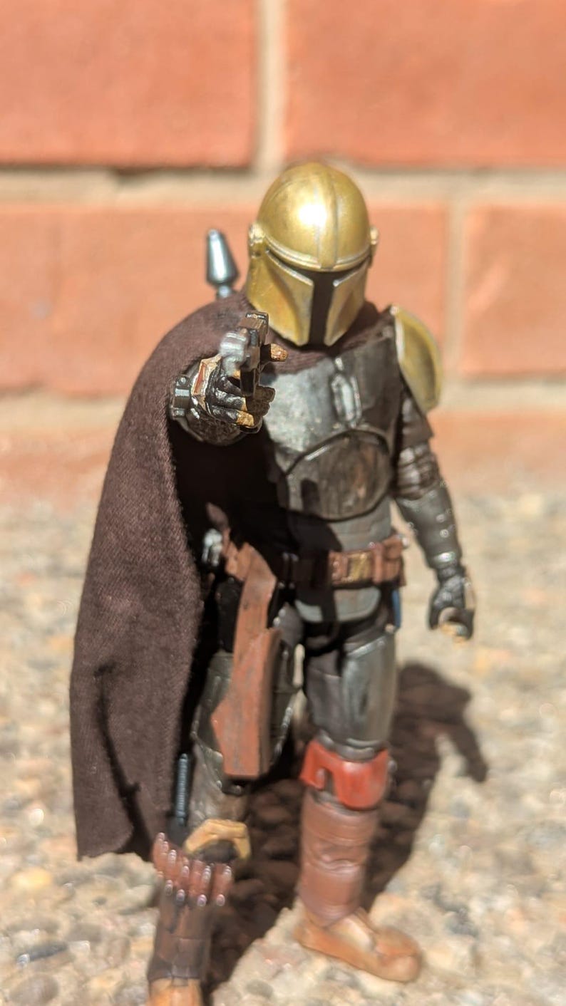 K&ouml;nnte beinhalten: Detaillierte Actionfigur eines Charakters in metallischer R&uuml;stung, mit goldenem Helm und braunem Umhang. Die Figur h&auml;lt eine Waffe und posiert vor einem Hintergrund aus Backsteinmauer. Die Figur stammt aus dem Star Wars-Franchise.