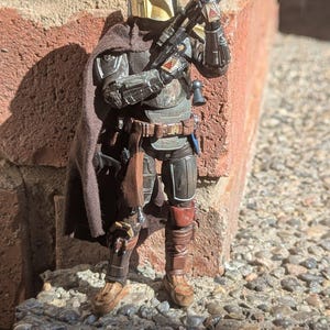 K&ouml;nnte beinhalten: Detaillierte Actionfigur eines mandalorianischen Kriegers in dunkler R&uuml;stung mit goldenem Helm. Die Figur h&auml;lt ein Blastergewehr, steht auf einer Steinoberfl&auml;che und lehnt an einer Backsteinmauer. Die Figur hat einen braunen Umhang und diverses Zubeh&ouml;r.