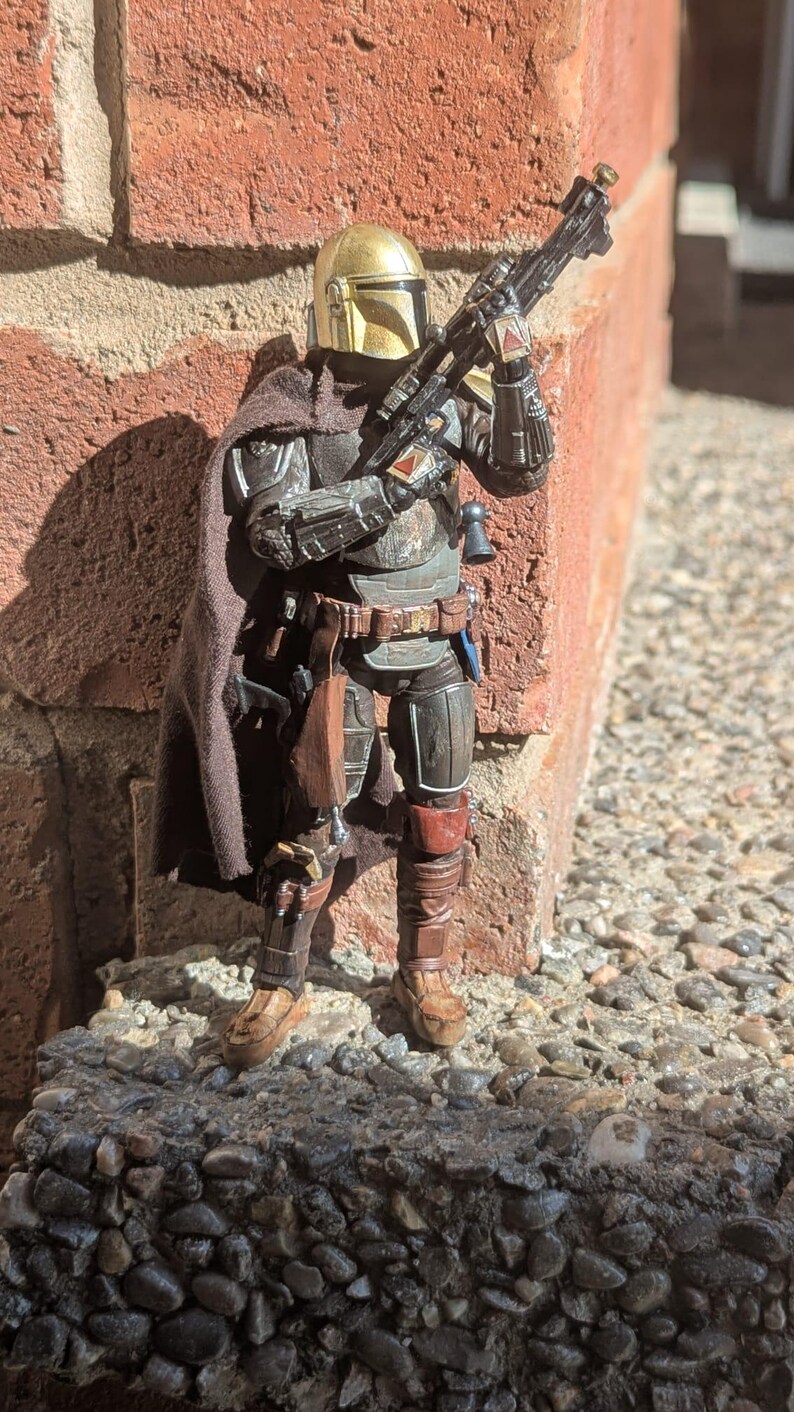 K&ouml;nnte beinhalten: Detaillierte Actionfigur eines mandalorianischen Kriegers in dunkler R&uuml;stung mit goldenem Helm. Die Figur h&auml;lt ein Blastergewehr, steht auf einer Steinoberfl&auml;che und lehnt an einer Backsteinmauer. Die Figur hat einen braunen Umhang und diverses Zubeh&ouml;r.