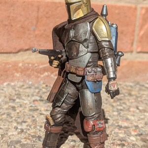 K&ouml;nnte beinhalten: Detaillierte Actionfigur einer Figur mit goldenem Helm und R&uuml;stung, die eine Blasterpistole h&auml;lt. Die Figur hat einen braunen Umhang, einen blauen Jetpack und posiert vor einer Backsteinmauer.
