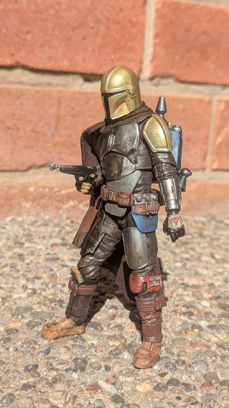 K&ouml;nnte beinhalten: Detaillierte Actionfigur einer Figur mit goldenem Helm und R&uuml;stung, die eine Blasterpistole h&auml;lt. Die Figur hat einen braunen Umhang, einen blauen Jetpack und posiert vor einer Backsteinmauer.
