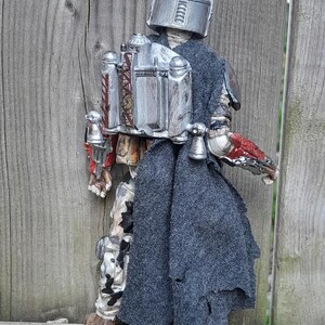 K&ouml;nnte beinhalten: Eine silberne und graue Actionfigur eines mandalorianischen Kriegers, der einen Helm und einen zerfetzten grauen Umhang tr&auml;gt. Die Figur h&auml;lt ein Blastergewehr in der rechten Hand und hat einen Jetpack auf dem R&uuml;cken.