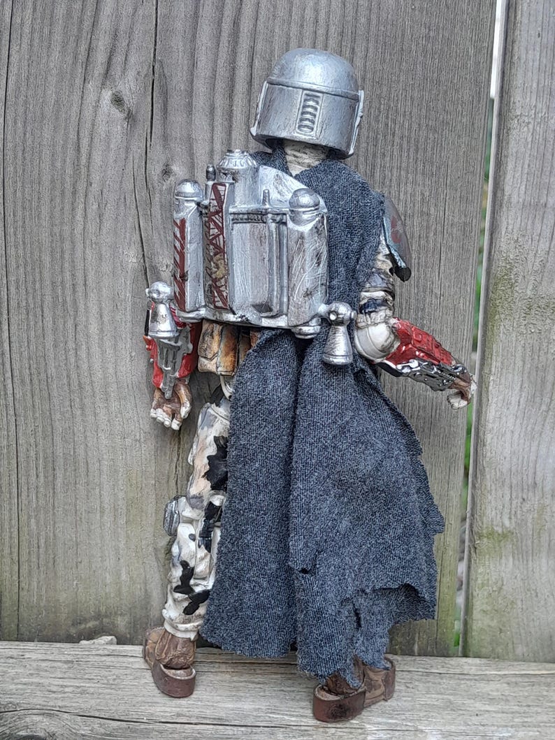 K&ouml;nnte beinhalten: Eine silberne und graue Actionfigur eines mandalorianischen Kriegers, der einen Helm und einen zerfetzten grauen Umhang tr&auml;gt. Die Figur h&auml;lt ein Blastergewehr in der rechten Hand und hat einen Jetpack auf dem R&uuml;cken.