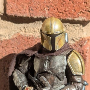 K&ouml;nnte beinhalten: Detaillierte Actionfigur eines Charakters mit goldenem und silbernem Helm und R&uuml;stung, der ein Blaster-Gewehr h&auml;lt. Die Figur hat einen braunen Umhang und steht vor einer Backsteinmauer.