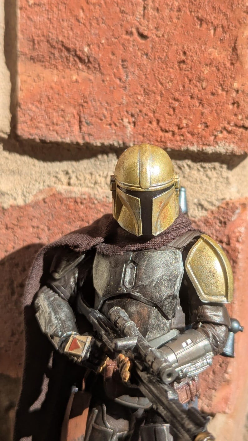 K&ouml;nnte beinhalten: Detaillierte Actionfigur eines Charakters mit goldenem und silbernem Helm und R&uuml;stung, der ein Blaster-Gewehr h&auml;lt. Die Figur hat einen braunen Umhang und steht vor einer Backsteinmauer.