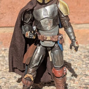 K&ouml;nnte beinhalten: Detaillierte Actionfigur eines Charakters in metallischer R&uuml;stung, mit goldenem Helm und Schulterpolstern. Die Figur hat einen braunen Umhang und G&uuml;rtel, mit einer Blasterpistole. Die Figur posiert vor einer Backsteinmauer.