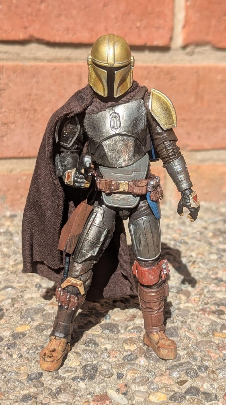 K&ouml;nnte beinhalten: Detaillierte Actionfigur eines Charakters in metallischer R&uuml;stung, mit goldenem Helm und Schulterpolstern. Die Figur hat einen braunen Umhang und G&uuml;rtel, mit einer Blasterpistole. Die Figur posiert vor einer Backsteinmauer.
