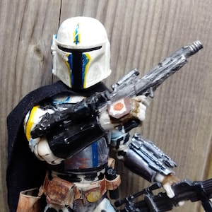 May include: A detailed action figure of a Mandalorian warrior, featuring a white helmet with blue and yellow accents. The figure is clad in armor with a black cape and holds two silver blasters. The figure has a brown belt and holster.