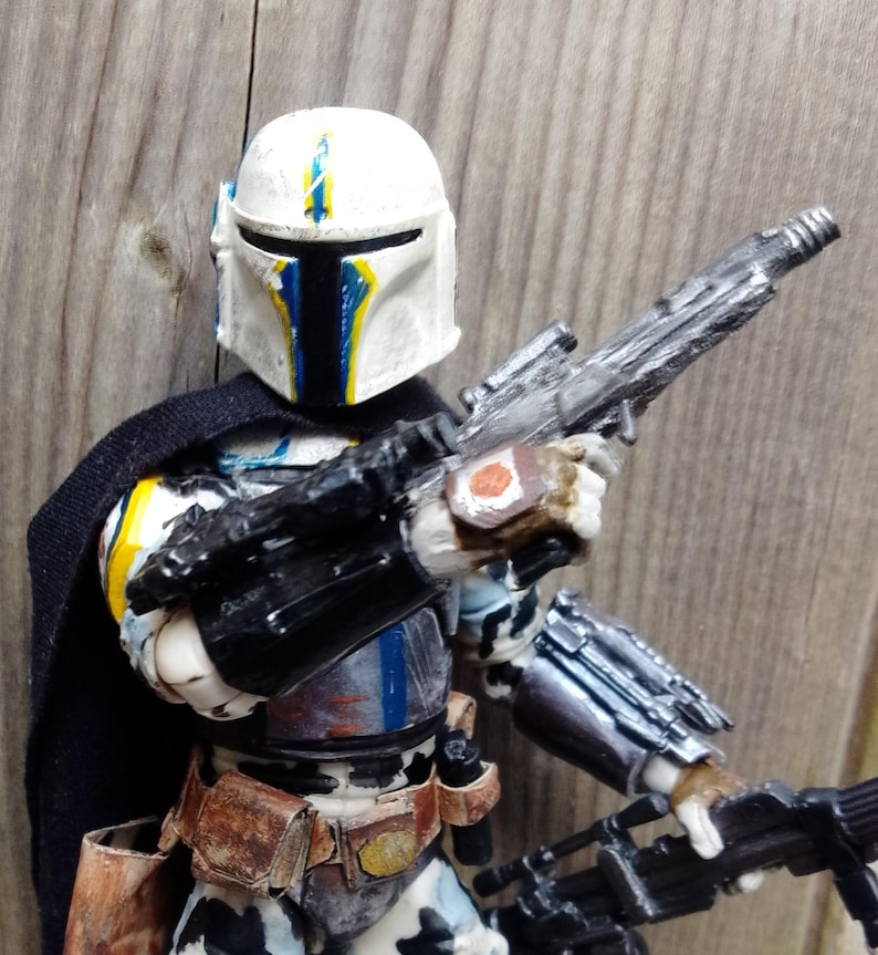 May include: A detailed action figure of a Mandalorian warrior, featuring a white helmet with blue and yellow accents. The figure is clad in armor with a black cape and holds two silver blasters. The figure has a brown belt and holster.