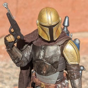 K&ouml;nnte beinhalten: Detaillierte Actionfigur eines Charakters mit goldenem Helm und R&uuml;stung, der eine schwarze Blaster-Pistole h&auml;lt. Die Figur hat einen braunen Umhang und einen Werkzeugg&uuml;rtel mit Taschen. Die R&uuml;stung hat ein verwittertes Aussehen mit Gold-, Silber- und Braunt&ouml;nen.