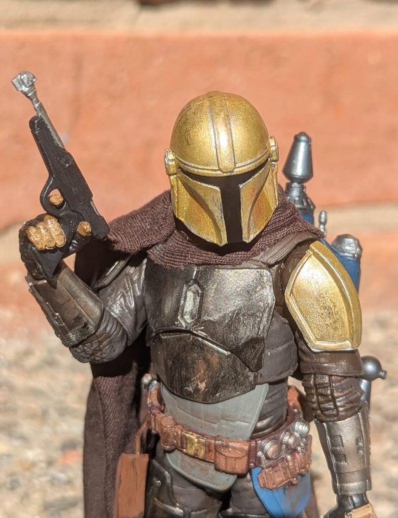 K&ouml;nnte beinhalten: Detaillierte Actionfigur eines Charakters mit goldenem Helm und R&uuml;stung, der eine schwarze Blaster-Pistole h&auml;lt. Die Figur hat einen braunen Umhang und einen Werkzeugg&uuml;rtel mit Taschen. Die R&uuml;stung hat ein verwittertes Aussehen mit Gold-, Silber- und Braunt&ouml;nen.