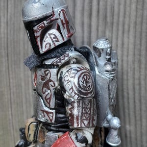 K&ouml;nnte beinhalten: Eine silberne und rote Actionfigur eines mandalorianischen Kriegers mit einem Jetpack und einem Blastergewehr. Die Figur ist detailliert und hat einen verwitterten Look.