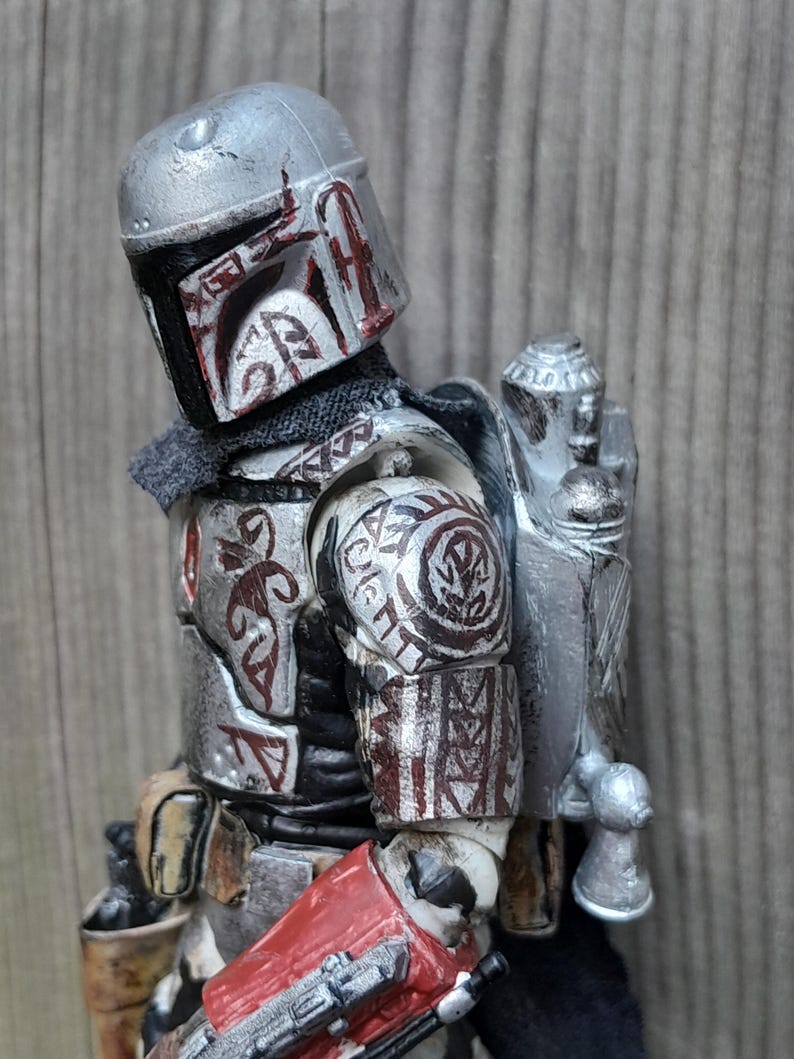 K&ouml;nnte beinhalten: Eine silberne und rote Actionfigur eines mandalorianischen Kriegers mit einem Jetpack und einem Blastergewehr. Die Figur ist detailliert und hat einen verwitterten Look.