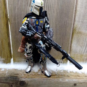 May include: A detailed action figure of a character in a white, blue, and black armored suit, holding a large black rifle. The figure has a helmet with yellow and blue accents and silver boots. The figure is standing in front of a wooden fence.