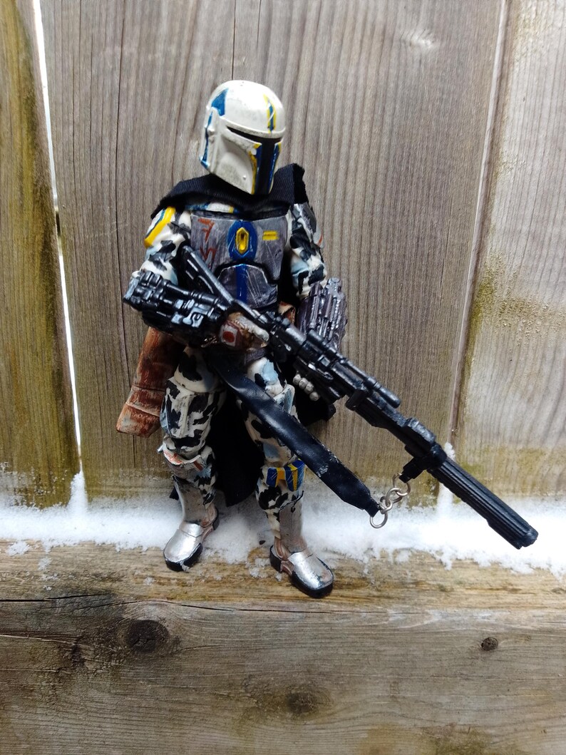 May include: A detailed action figure of a character in a white, blue, and black armored suit, holding a large black rifle. The figure has a helmet with yellow and blue accents and silver boots. The figure is standing in front of a wooden fence.