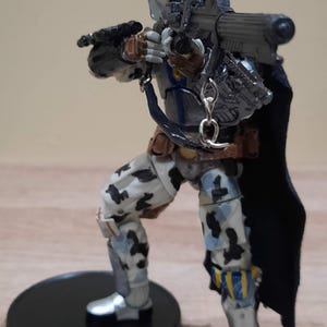 May include: A detailed action figure of a character in a white helmet with blue and yellow accents, holding a blaster rifle. The figure wears armor with camouflage patterns and a black cape, standing on a black circular base.