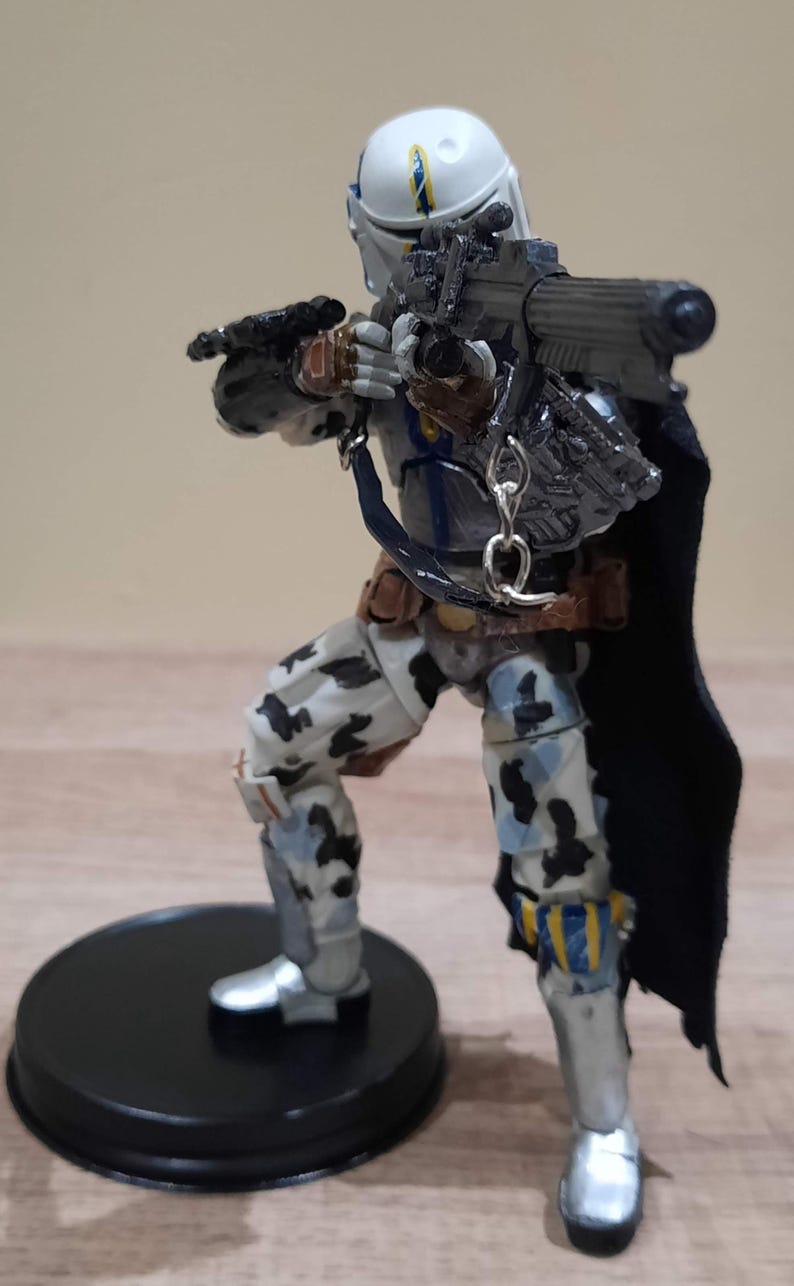 May include: A detailed action figure of a character in a white helmet with blue and yellow accents, holding a blaster rifle. The figure wears armor with camouflage patterns and a black cape, standing on a black circular base.
