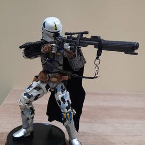 May include: A detailed action figure of a character in a white and blue helmet, holding a black rifle. The figure is wearing a black cape, brown belt, and gray and white patterned armor. The figure is standing on a black circular base.