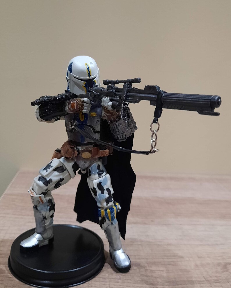 May include: A detailed action figure of a character in a white and blue helmet, holding a black rifle. The figure is wearing a black cape, brown belt, and gray and white patterned armor. The figure is standing on a black circular base.