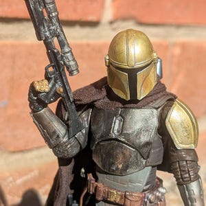 K&ouml;nnte beinhalten: Detaillierte Actionfigur einer Figur aus einem Science-Fiction-Franchise. Die Figur ist in metallischer R&uuml;stung mit einem goldenen Helm gekleidet und h&auml;lt ein schwarzes Gewehr. Die Figur hat einen braunen Umhang und einen Werkzeugg&uuml;rtel.