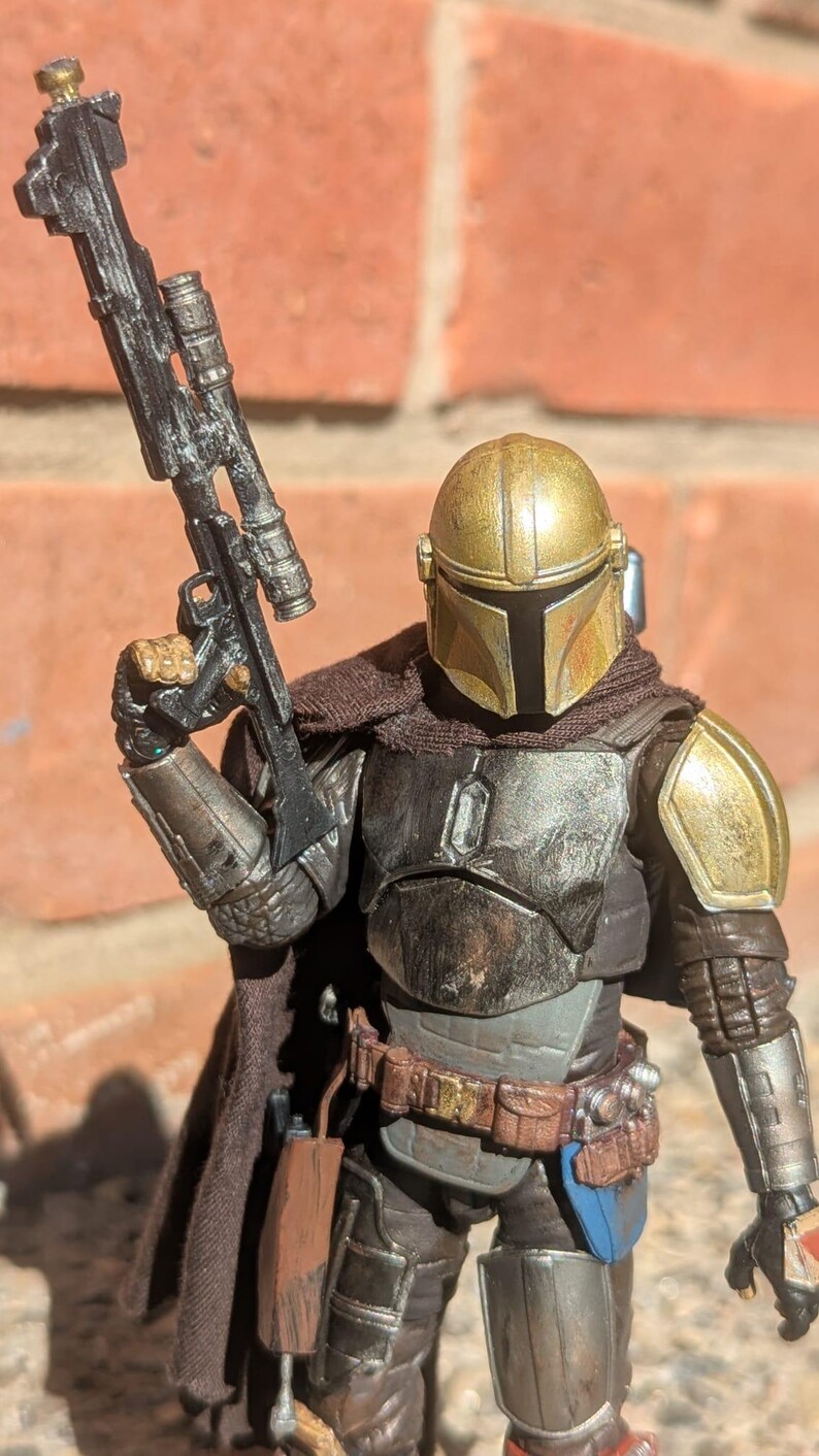 K&ouml;nnte beinhalten: Detaillierte Actionfigur einer Figur aus einem Science-Fiction-Franchise. Die Figur ist in metallischer R&uuml;stung mit einem goldenen Helm gekleidet und h&auml;lt ein schwarzes Gewehr. Die Figur hat einen braunen Umhang und einen Werkzeugg&uuml;rtel.