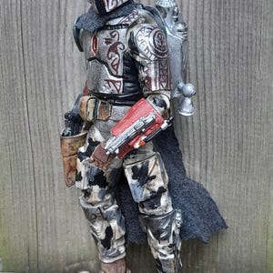 K&ouml;nnte beinhalten: Eine silberne und rote Actionfigur eines Mandalorianer-Kriegers mit einem Jetpack und einem Tarnmuster an den Beinen. Die Figur hat ein detailliertes Design mit Stammesmarkierungen und einem schwarzen Umhang.