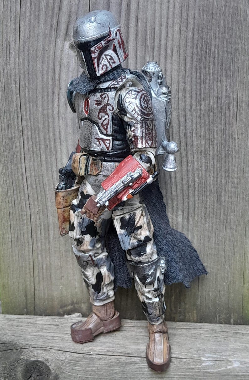 K&ouml;nnte beinhalten: Eine silberne und rote Actionfigur eines Mandalorianer-Kriegers mit einem Jetpack und einem Tarnmuster an den Beinen. Die Figur hat ein detailliertes Design mit Stammesmarkierungen und einem schwarzen Umhang.