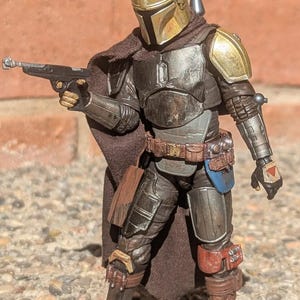 K&ouml;nnte beinhalten: Detaillierte Actionfigur eines mandalorianischen Kriegers in abgenutzter R&uuml;stung. Die Figur hat einen goldenen Helm, Schulterpolster und eine Blasterpistole. Die Figur ist mit einem Blaster in der Hand in Kampfpose. Die Figur ist etwa 15 cm gro&szlig;.