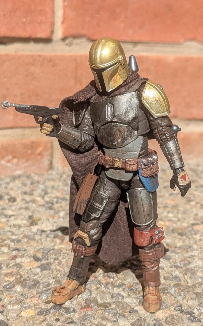K&ouml;nnte beinhalten: Detaillierte Actionfigur eines mandalorianischen Kriegers in abgenutzter R&uuml;stung. Die Figur hat einen goldenen Helm, Schulterpolster und eine Blasterpistole. Die Figur ist mit einem Blaster in der Hand in Kampfpose. Die Figur ist etwa 15 cm gro&szlig;.
