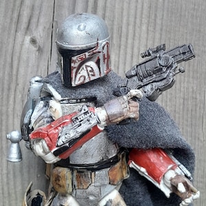 K&ouml;nnte beinhalten: Eine abgenutzte Actionfigur eines Kopfgeldj&auml;gers mit einem silbernen und grauen Helm mit roten Markierungen, einem grauen Umhang und einem schwarzen Blaster.