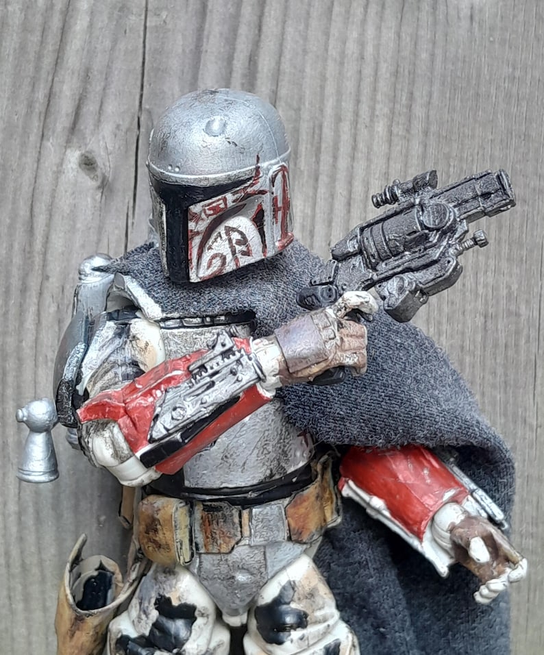 K&ouml;nnte beinhalten: Eine abgenutzte Actionfigur eines Kopfgeldj&auml;gers mit einem silbernen und grauen Helm mit roten Markierungen, einem grauen Umhang und einem schwarzen Blaster.