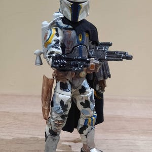 May include: A detailed action figure of a character in a white, blue, and gray armored suit, holding a black rifle. The figure has a black cape and a jetpack. The helmet is white with blue accents. The figure is standing on a wooden surface.