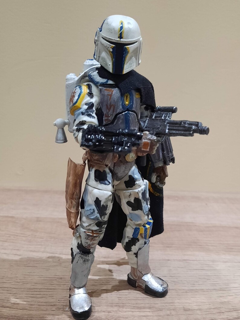 May include: A detailed action figure of a character in a white, blue, and gray armored suit, holding a black rifle. The figure has a black cape and a jetpack. The helmet is white with blue accents. The figure is standing on a wooden surface.